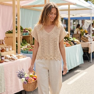 May include: A beige short-sleeved crochet top with a V-neckline and intricate vertical lace patterns, paired with light-colored trousers. The top features a scalloped hemline and is crafted with a delicate, open-work knit design throughout.