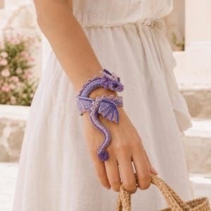 Op de afbeelding: Een gehaakte drakenarmband in paars en licht lavendel die om de pols wikkelt. De draak heeft een gedetailleerde kop met kleine oortjes, een lang gebogen lichaam en een staart die aan het uiteinde krult, gemaakt met fijn garen en steken.