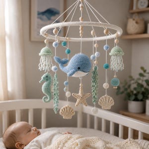 May include: A nursery mobile featuring crocheted sea creatures including a blue whale, a seahorse, a starfish, two jellyfish, and two seashells. The items hang from a white circular frame with wooden beads and small blue and white spheres.