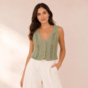 May include: A sage green sleeveless crochet waistcoat featuring a V-neckline, a vertical open-knit pattern, and a front button closure with five small light-coloured circular buttons. The garment is paired with high-waisted off-white trousers.