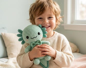 May include: A light mint green crocheted axolotl plush toy featuring a small bow tie at the neck, black circular eyes, and pink embroidered cheek details. The stuffed creature has three feathery external gills on each side of its round head.