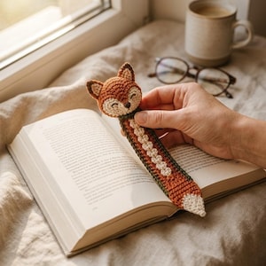 May include: A crocheted fox bookmark featuring a rust-orange head with dark brown ear tips, a cream-colored face, and a long, flat body with a central cream-colored stripe and a white tail tip, resting between the pages of an open book.