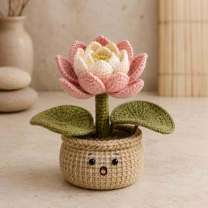 Puede incluir: Una flor de loto de ganchillo con pétalos rosas y crema y un tallo verde, con dos hojas verdes. La flor está plantada en una maceta de ganchillo color canela con un borde trenzado y una cara bordada con dos ojos negros y una boca abierta.
