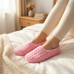 May include: A pair of light pink slippers featuring a textured, crocheted knit pattern. The slippers have a rounded toe design and a slip-on style, covering the foot up to the ankle area with a consistent, dense yarn construction throughout.