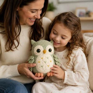 May include: A sage green and cream crocheted owl plush toy with large dark eyes, a small yellow beak, and orange feet. The owl features a textured stitch pattern on its body with small embroidered floral accents in green and yellow thread.