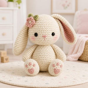 May include: A cream-colored crochet bunny plush toy featuring long ears with pink inner lining, black safety eyes, a pink stitched nose, and rosy cheeks. A small pink flower with green leaves is attached to the side of the head, and the feet have pink heart-shaped pads.