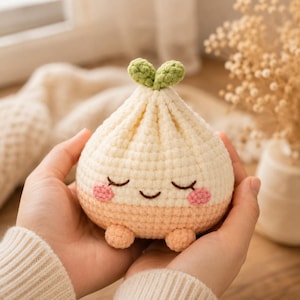 May include: A round, cream-colored crochet plush toy shaped like a dumpling with a small green sprout on top. It features embroidered closed eyes, a curved smile, and two small pink circular cheeks. The base is a light peach color with two small feet.