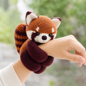 May include: A crocheted red panda plush toy featuring a brown and white face with black eyes and a dark nose. The body is made of brown yarn with dark maroon limbs and a striped tail, designed to wrap around an arm with its long, flexible arms.