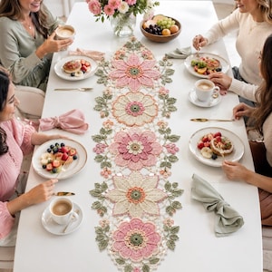 Può includere: Un lungo runner da tavola rettangolare all'uncinetto con un motivo floreale ripetuto composto da grandi fiori a più strati nelle tonalità del rosa, crema e verde salvia. L'intricato design in pizzo include petali e foglie dettagliati uniti da una rete.