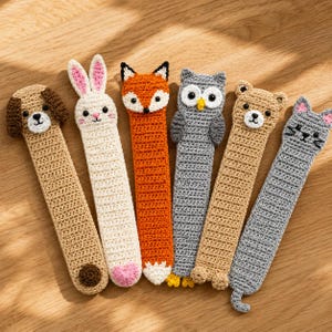 May include: A set of six crochet animal bookmarks featuring a tan dog, a white rabbit, an orange fox, a gray owl, a light brown bear, and a gray cat. Each bookmark has a long, flat, rectangular body with a detailed animal head at the top.