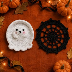 May include: A white crocheted ghost coaster with black bead eyes and pink stitched cheeks, positioned next to a black crocheted spider web coaster. Both items are circular and feature intricate yarn work, displayed on a textured orange surface.