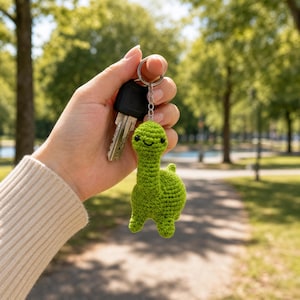 May include: A small, bright green crocheted dinosaur keychain featuring a long neck, a rounded body, and two small black bead eyes with a stitched smile. The dinosaur is attached to a silver-toned metal split ring that holds a single metal key.