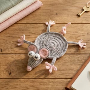 May include: A gray crocheted coaster shaped like a flat mouse with a round body, four splayed limbs, a curled tail, and two large ears with pink centers. The mouse has a pointed snout with a pink nose and two white eyes featuring black X stitches.