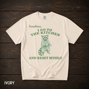 Könnte beinhalten: Ein elfenbeinfarbenes Kurzarm-T-Shirt mit einer grünen Grafik eines stehenden Waschbären in der Mitte. Der Text über dem Waschbären lautet "Sometimes, I GO TO THE KITCHEN" und der Text darunter "AND RESET MYSELF" in einer grünen Serifenschrift.