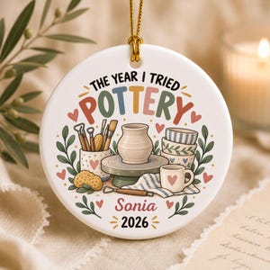 May include: A round white ceramic ornament featuring the text THE YEAR I TRIED POTTERY, Sonia, and 2026. The illustration shows a pottery wheel, a clay pot, stacked bowls, a mug, and tools, decorated with colorful hearts and green leaves.
