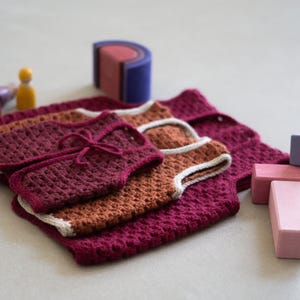 May include: Three crocheted waistcoats in burgundy and rust orange, featuring a textured stitch pattern and cream-coloured trim. The waistcoats are layered together, with the top piece secured by a matching yarn tie, accompanied by small wooden toy figures and blocks.