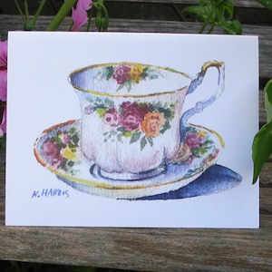 May include: A watercolor painting of a teacup and saucer with a floral pattern. The cup is white with pink, yellow, and red flowers. The saucer is white with a pink, yellow, and green floral pattern. The artist's signature, "K. Harris", is in the bottom left corner.
