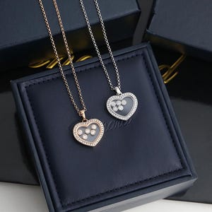 May include: Two heart-shaped pendant necklaces, one in rose gold and one in silver-toned metal. Each pendant features a clear centre with five small, round, bezel-set stones that move freely, surrounded by a border of tiny pavé-set stones.