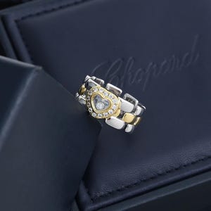 May include: A two-tone ring featuring a central heart-shaped motif outlined with small round diamonds and a single floating diamond at its centre. The band is constructed from alternating silver-toned and gold-toned polished metal links.