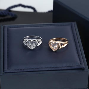 Könnte beinhalten: Zwei Ringe mit einem herzförmigen Rahmen, in dem sich drei bewegliche runde Diamanten befinden. Ein Ring ist aus poliertem silberfarbenem Metall gefertigt, der andere aus poliertem roségoldfarbenem Metall, beide mit geteilter Ringschiene.