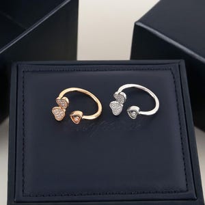 May include: Two open-style rings, one in gold-tone and one in silver-tone metal. Each ring features two heart-shaped motifs at the ends, with one heart encrusted with small clear stones and the other containing a single floating stone inside a frame.
