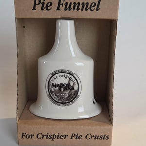 May include: A white ceramic pie funnel with a bell-shaped base and a narrow top, housed in a brown cardboard box. The box is printed with the text Pie Funnel at the top and For Crispier Pie Crusts at the bottom. A circular logo is on the funnel.