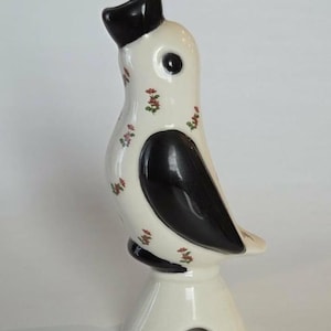 May include: A ceramic bird figurine featuring a glossy white body with black accents on the head, wing, and base. The white surface is decorated with a repeating pattern of small red and green floral sprigs. The bird stands on a pedestal base.