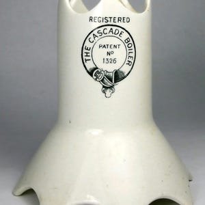 May include: A white ceramic boiler component with a flared, scalloped base and a crown-like top edge. The front features a black printed emblem with the text REGISTERED THE CASCADE BOILER PATENT NO 1326 inside a circular belt-style design.