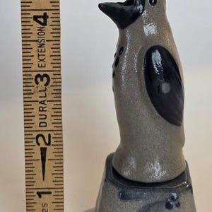 May include: A ceramic penguin figurine with a speckled gray body and dark blue accents on the wings, beak, and base. A wooden ruler placed next to the figurine shows it stands approximately 5.5 inches tall, featuring a hand-painted blue floral design.