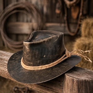 Op de afbeelding: Een donkerbruine, verweerde cowboyhoed met een doorleefde afwerking en een ingedeukte kroon. De hoed is voorzien van een lichtbruine, gevlochten touwband die rond de basis van de kroon loopt en aan de rechterkant in een knoop zit.