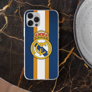 May include: A rectangular smartphone case featuring a textured blue background with a vertical white stripe flanked by two thin gold stripes. Centered on the case is the Real Madrid football club crest, displaying a gold crown above a blue and gold logo.