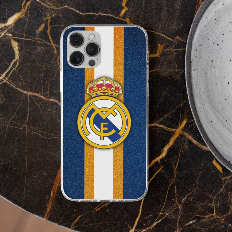 May include: A rectangular smartphone case featuring a textured blue background with a vertical white stripe flanked by two thin gold stripes. Centered on the case is the Real Madrid football club crest, displaying a gold crown above a blue and gold logo.