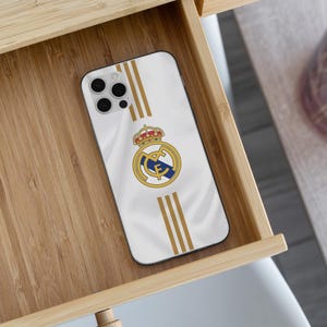 May include: A white smartphone case featuring the Real Madrid football club crest in the center, flanked by three vertical gold stripes above and below the logo. The case has a cutout for a triple-lens camera system on the upper left corner.