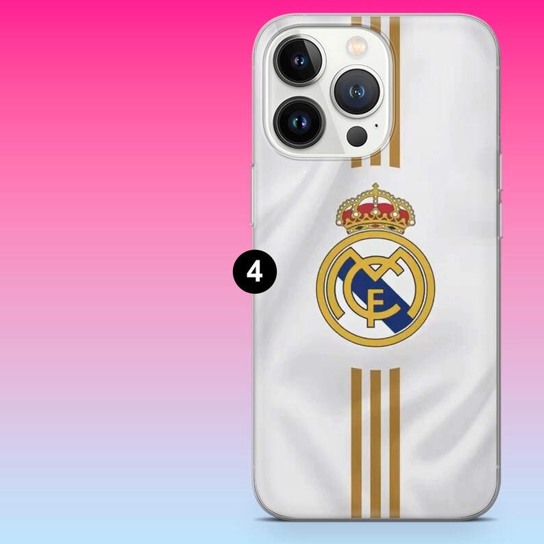 May include: A white smartphone case featuring the Real Madrid football club logo in gold, blue, and red, centered vertically. Three vertical gold stripes extend above and below the crest. The case has a cutout for a triple-lens camera system.