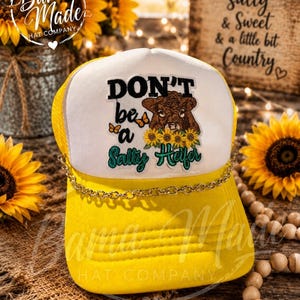 Peut inclure: Une casquette de camionneur jaune vif avec un panneau avant blanc orné d'un motif représentant une vache brune, des tournesols et des papillons. Le texte "DON'T be a Salty Heifer" est inscrit en noir et bleu sarcelle. Une chaîne dorée orne la visière.