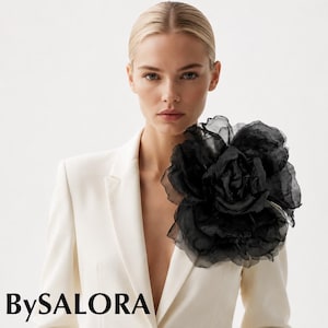 Peut inclure: Un blazer couleur crème orné d'une grande fleur en tissu noir à plusieurs épaisseurs épinglée sur le revers gauche. Le blazer présente un col à revers et une fermeture à un bouton. Le texte BySALORA est imprimé en noir sur le côté gauche.