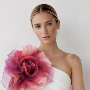 Peut inclure: Une robe blanche à une épaule ornée d'une grande fleur en organza superposée dans des tons rose, magenta et rouge foncé sur l'épaule. Le texte BySALORA est imprimé en noir sur le devant de la robe. Le tissu présente une texture froncée.