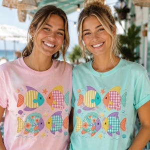 Puede incluir: Una camiseta rosa claro y una camiseta verde menta claro, cada una con un estampado gráfico de cuatro peces estilizados con varios patrones que incluyen rayas, cuadros vichy y flores, acentuados por pequeñas ilustraciones de estrellas de mar y conchas.