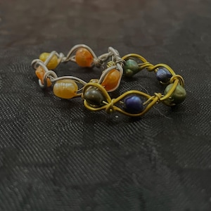 Può includere: Due anelli in filo metallico intrecciato con piccole perline irregolari. Un anello è realizzato in filo argentato con perline arancioni e gialle, mentre il secondo anello è in filo dorato con perline blu scuro, verdi e viola.