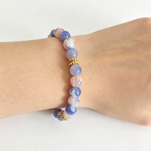 Könnte beinhalten: Ein Perlenarmband mit runden, polierten Steinen in Blau-, Weiß- und zarten Rosatönen. Die Perlen sind in einer durchgehenden Schlaufe angeordnet und mit zwei kleinen, strukturierten, goldfarbenen Metallzwischenperlen versehen.