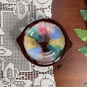 May include: A brown ceramic dish with a colorful, abstract design in the center. The colors include pink, blue, yellow, and green. The dish has two small handles and is set on a white lace doily.