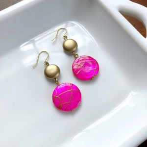 May include: A pair of dangle earrings featuring a gold-tone metal and bright pink, round, beaded accents. The pink beads have a gold-colored, web-like design. The earrings are displayed on a white, rectangular dish.