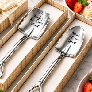 May include: Two silver-toned metal spoons shaped like miniature shovels, each featuring a triangular handle and a bowl engraved with the text "Paige's Ice Cream Shovel". One spoon bowl also includes a small graphic of an ice cream sundae.
