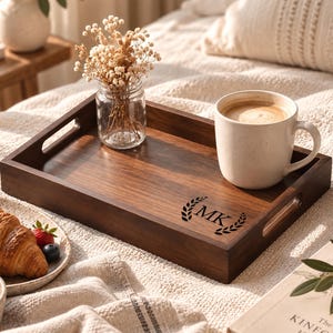 May include: A rectangular dark brown wooden serving tray with cutout handles, featuring a laser-engraved laurel wreath design surrounding the letters MK. The tray holds a small glass jar with dried flowers and a speckled off-white ceramic mug.