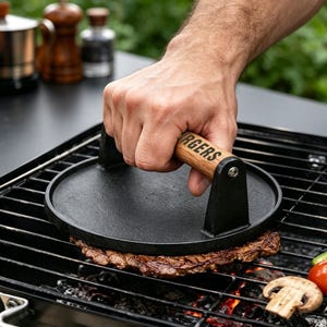 May include: A round black cast iron grill press with a wooden handle featuring the text "BURGERS" printed in black. The heavy-duty tool is positioned on top of a piece of meat cooking on a metal barbecue grill grate over hot glowing coals.