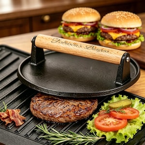 May include: A round black cast iron burger press with a light wood handle featuring the engraved text Johnson's Burgers. The press sits on a black ridged grill pan next to a cooked beef patty, lettuce, tomato slices, a pickle, and bacon.