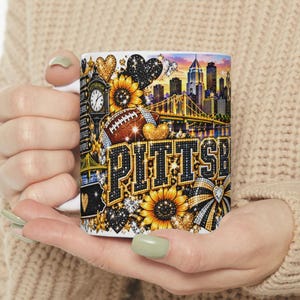 Peut inclure: Un mug en céramique blanche orné d'un imprimé graphique vibrant représentant les gratte-ciel de Pittsburgh, un ballon de football, des tournesols et des cœurs dans des tons noir et or. Le mot PITTSBURGH est écrit en grandes lettres dorées.