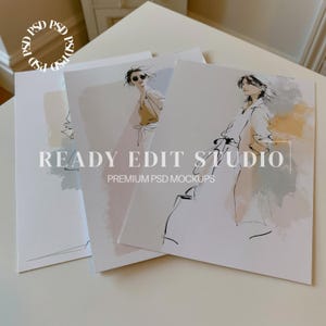 May include: Three stacked rectangular paper prints featuring fashion illustrations with watercolor-style washes in beige, gray, and white. The prints display line drawings of figures in clothing, with the text READY EDIT STUDIO PREMIUM PSD MOCKUPS.