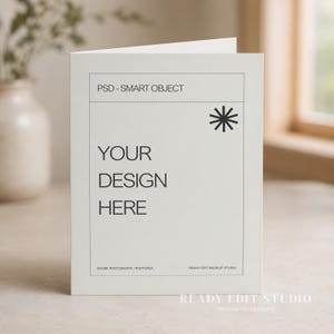 May include: A white folded greeting card mockup featuring a rectangular frame with the text PSD - SMART OBJECT at the top and YOUR DESIGN HERE in the center. A black star-like icon is in the upper right corner, with small text at the bottom.