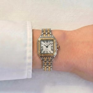 May include: A two-tone wristwatch featuring a square silver-tone case with a gold-tone bezel. The white dial displays black Roman numeral hour markers and the text GREELY. The bracelet consists of alternating silver and gold-tone metal links.